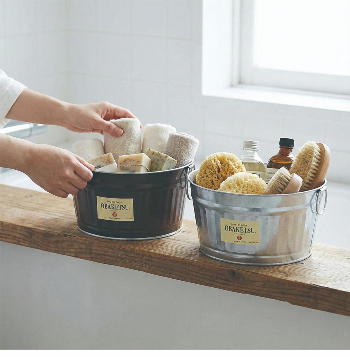 兩個復古風格的 OBAKETSU 桶，一個深棕色，一個銀色，裝滿了毛巾、肥皂、海綿和刷子，放在木製檯面上。