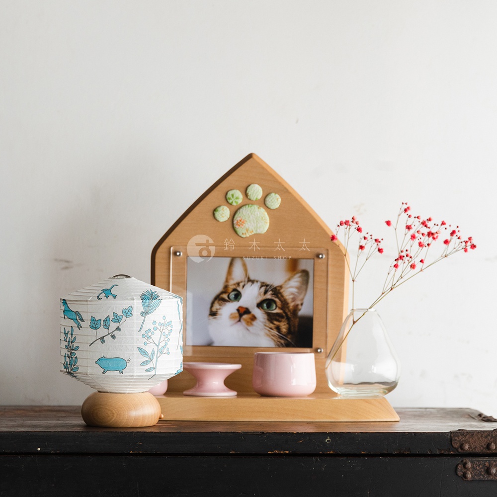 白色紙燈籠，帶有藍色植物和豬的圖案，底部為木製底座，旁邊是一個木製相框，內有貓咪照片，相框頂部裝飾有綠色爪印。