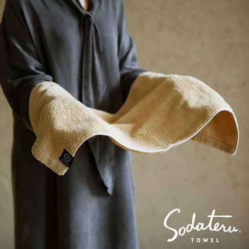 一件深灰色的長袍，雙手捧著一條淺黃色的毛巾，毛巾上有一個黑色標籤，下方印有 Sodateru TOWEL 的白色文字。