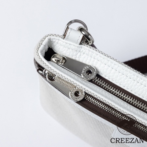 這是一張由上往下的近攝視角照片，展示了 CREEZAN 品牌隨身斜背包的細節。包身主體由米白色編織帆布材質製成，上方配有白色荔枝紋皮革包邊。頂部設有兩層拉鍊開口，拉鍊頭選用銀色金屬材質，並刻有品牌名稱。包包一側連接著銀色金屬扣環，整體呈現出簡約精緻的質感。