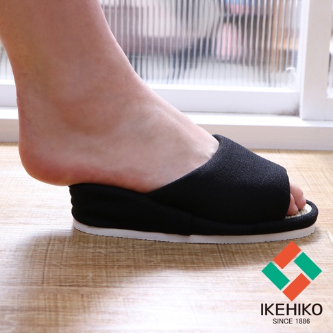 黑色藺草編織面料的室內拖鞋，鞋面為開放式設計，露出腳趾。鞋床為厚實的坡跟楔形，外底為白色，與木質地板形成對比。