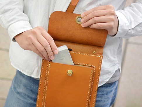 一個手持著棕色皮革厚實單肩包（CP-61），將手機放入包包的外部口袋。包包採用手工縫製，附有金屬扣和金屬孔眼。