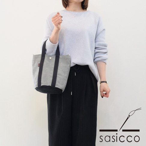 一位女士手提一個三河木綿刺子織兩用水桶包，包身為黑白棋盤格圖案，配有黑色提把與底部，整體風格簡潔。