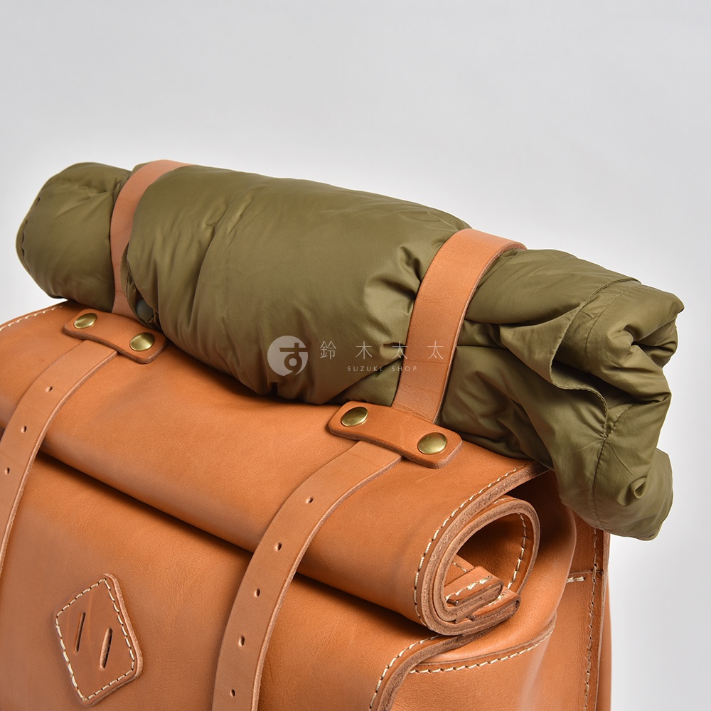 職人手作的棕色皮革後背包，頂部捲起固定著軍綠色羽絨睡袋，以皮帶和金屬扣件牢固。