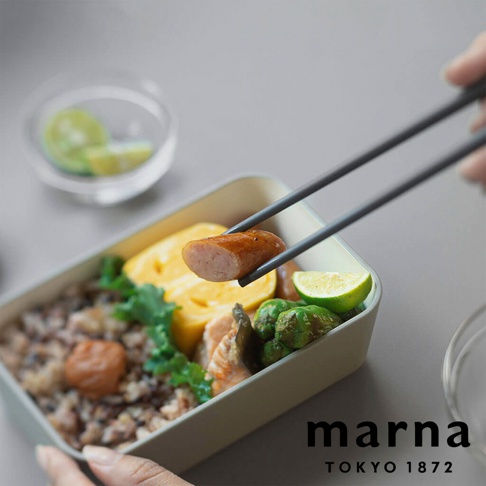 marna TOKYO 1872 品牌字樣搭配一雙筷子正在夾起午餐盒中的香腸，午餐盒內有米飯、玉子燒、鮭魚和蔬菜。