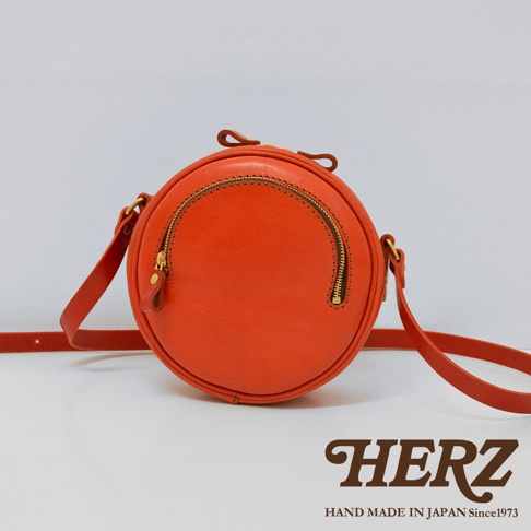 橘色皮革小圓包，圓形包身，正面有半圓形拉鍊開口，搭配金色拉鍊及同色系皮革拉頭，附可調式皮革背帶。