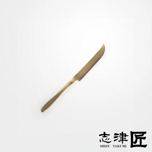 黃銅色起司刀，刀刃一側有鋸齒狀邊緣，刀柄圓潤，整體設計簡約，背景為純白色。