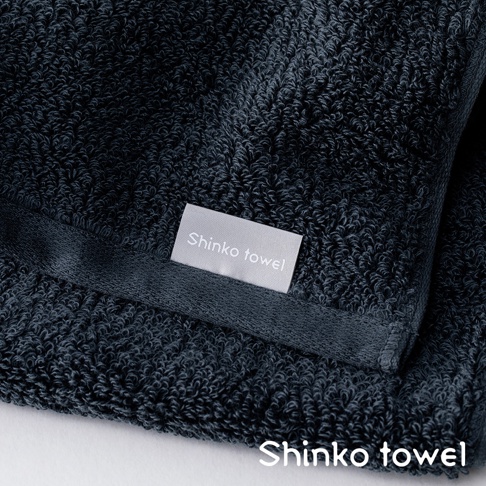 深藍色匹馬棉浴巾，帶有淺灰色標籤，標有「Shinko towel」字樣。浴巾材質柔軟，紋理細緻，呈現高品質觸感。