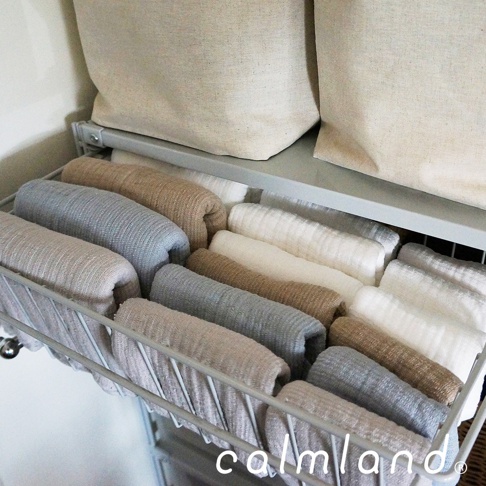 整齊摺疊的毛巾，顏色包含米白、淺灰、卡其色，材質呈現細緻織紋，擺放在金屬網籃中，上方另有兩個米白色布質收納袋。