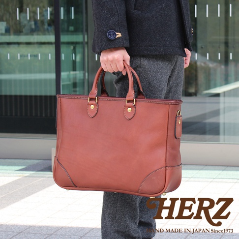 一位穿著深灰色大衣的男士正手提著一個日本HERZ品牌製作的棕色皮革商務托特包，該包款具有厚實的皮革質感與金屬扣件細節，整體展現出職人手作的簡約商務風格。