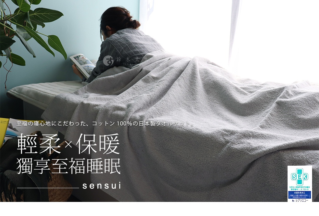 一條淺灰色、雙面毛圈織法的棉毛巾被，質地柔軟，蓋在一位正在閱讀的女性身上。