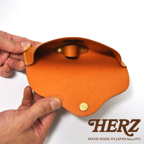 職人手作皮革眼鏡收納盒，橘棕色皮革，內部有固定帶與釦子，下方有品牌標誌HERZ HAND MADE IN JAPAN Since1973