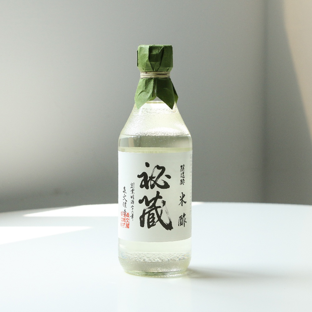 玻璃瓶裝的秘藏米醋，瓶身有白色標籤，上面有黑色的漢字「秘藏」及「米醋」字樣，瓶頸處以綠色皺紋紙包裹。