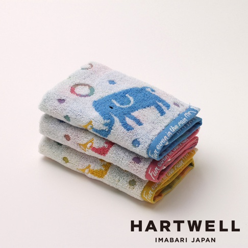 三條摺疊整齊的 Hartwell 今治毛巾由上至下依序為藍色、粉紅色與黃色，毛巾表面繡有藍色大象圖案以及小號角、圓圈與星星等可愛圖案，邊緣處印有 Fun even in the rain 的字樣。