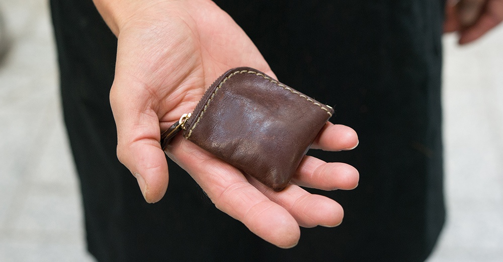 這是一張近距離特寫照片，一隻手掌心上放置著一個日本HERZ職人手作的棕色皮革小零錢包，零錢包呈現精巧的方型輪廓，外觀擁有柔軟的牛皮質感與經典的粗縫線手工細節，側邊配有方便開合的拉鍊設計。