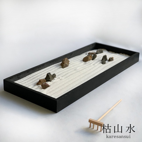 黑色長方形木製沙盤，內有細膩的白色沙子，耙出波浪紋理，散布著幾塊深淺不一的灰色、棕色石塊。右側有一迷你木製耙子，沙盤旁有「枯山水」中日文標示。