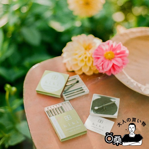 淺綠色包裝的香氛火柴禮盒，內含多根火柴，火柴頭為白色。旁邊還有一個裝有黑色圓形物和火柴的白色小盒。背景是模糊的黃色與粉色花朵及綠葉。