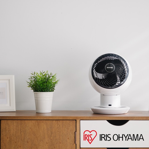白色圓形 Iris Ohyama PCF-SDC15T 空氣循環扇，配有黑色扇葉和底座，放置在木製家具上，旁邊有一盆綠色植物。
