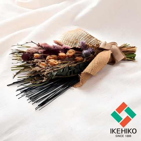 乾燥花束，包含紫色、粉色、米色、綠色等乾燥植物，搭配麻布裝飾，束綁於一側，底部為黑色細長條狀物。