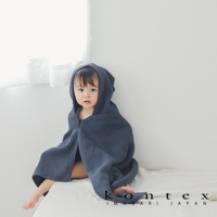 一名穿著深藍色針織純棉連帽浴巾的嬰兒，坐在白色背景前的床墊上。