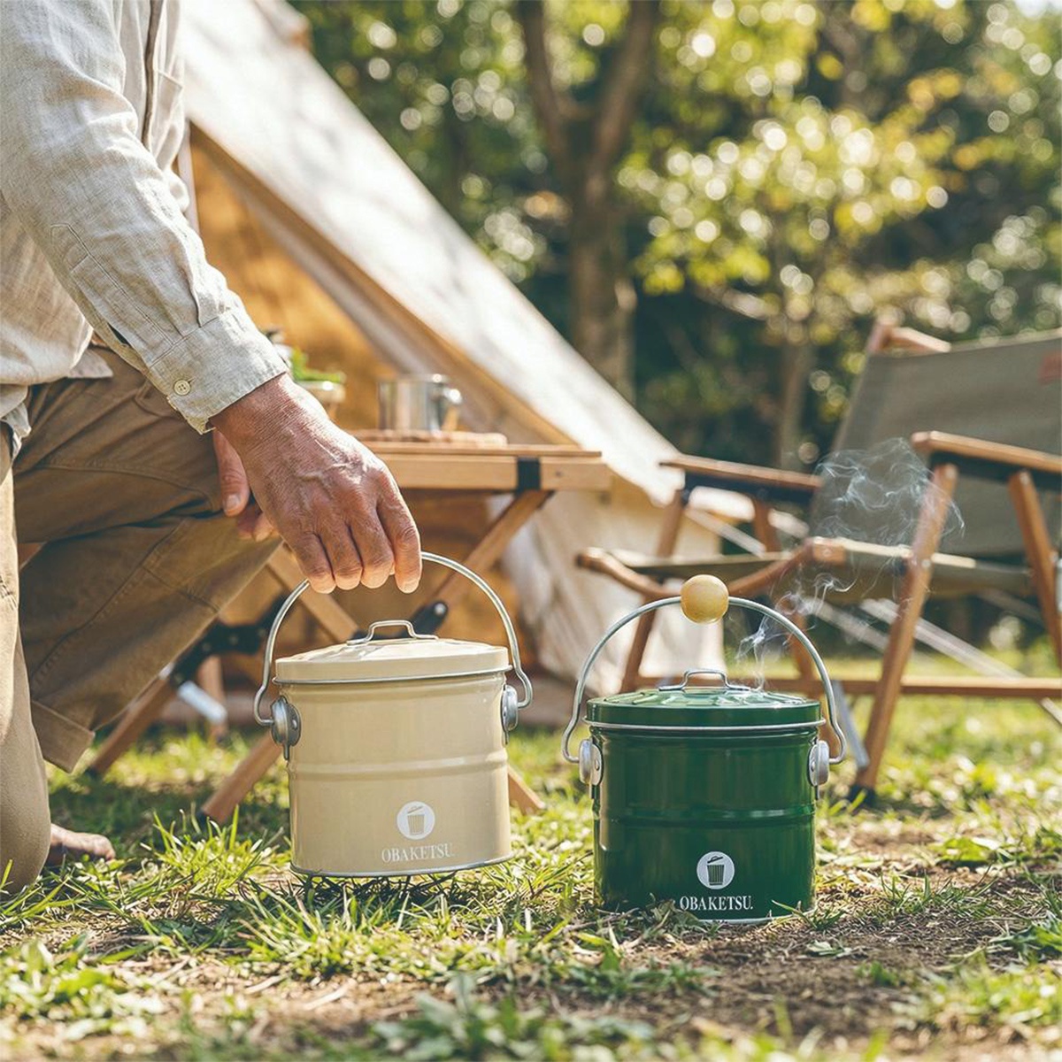 OBAKETSU 品牌的手工製小尺寸蚊香桶，一為米白色，另一為綠色，皆為圓筒形狀，附有金屬提把，置於草地上的露營場景中。