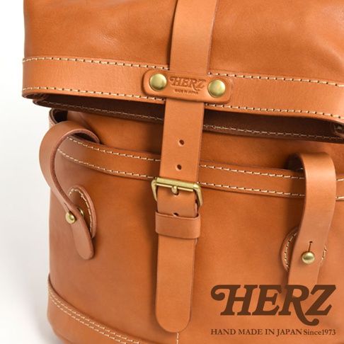 職人手作皮革雙口袋折蓋後背包，採用棕色皮革材質，配有黃銅釦環及針釦，車縫線清晰，展現精緻工藝。