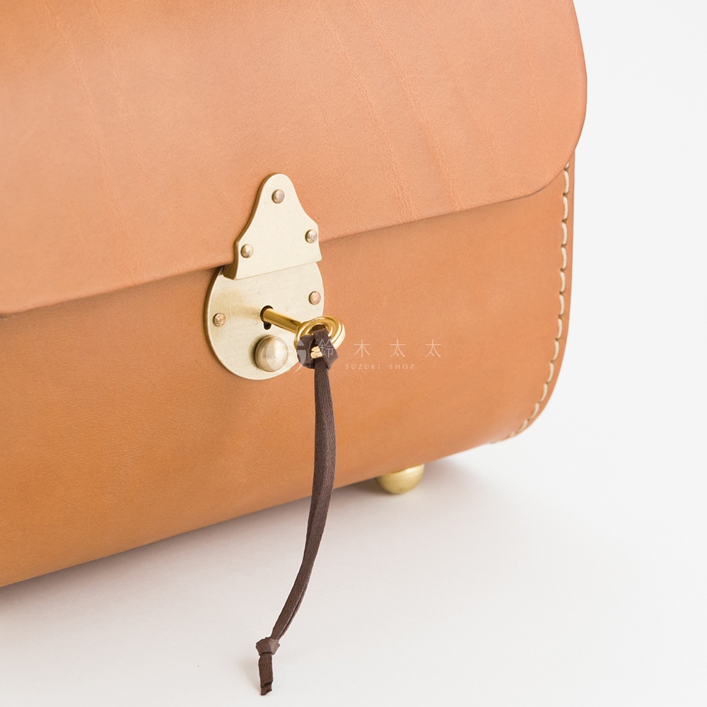 職人手作皮革復古杜勒斯手提包，採用棕褐色皮革，搭配黃銅色鎖頭、鑰匙及裝飾釘。包袋縫線清晰，鎖孔處懸掛一條深棕色皮革細繩，末端打結。