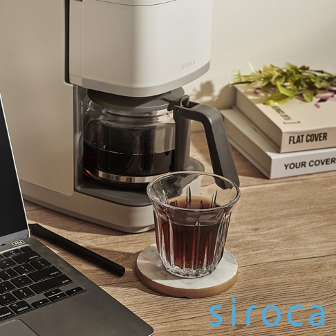 淺灰色的 siroca SC-C2510 石臼式全自動研磨咖啡機，帶有玻璃咖啡壺，壺中裝有深色咖啡。旁邊有一個玻璃杯，盛有咖啡，放在大理石杯墊上。右側是堆疊的書籍和綠色植物。左側有一個筆記型電腦。
