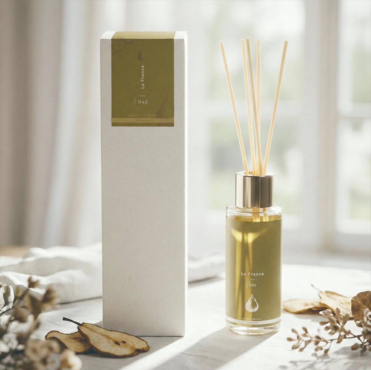 KYARA PERFUMERS La France 042 香氛擴香竹，搭配簡約的包裝盒，放置於布藝桌布上，周圍點綴著乾枯的梨子和枝葉，背景為柔和的窗戶光線。