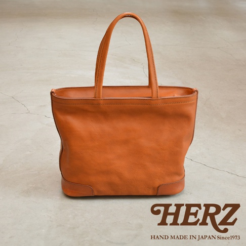 HERZ 品牌標誌，下方為 HAND MADE IN JAPAN Since1973 字樣，整體為一只橘棕色皮革托特包，包款線條簡潔，有兩個提把，底部有加厚的皮革裝飾。
