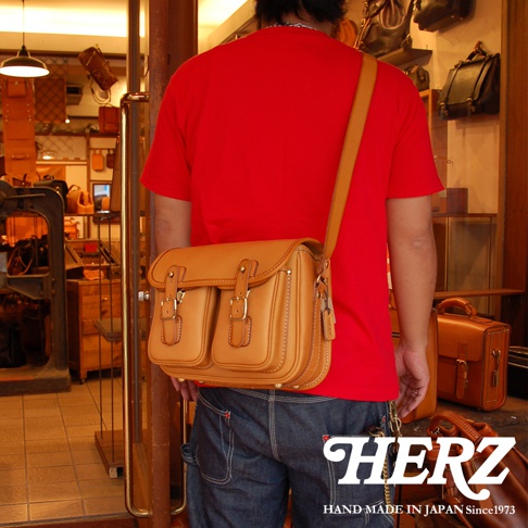 一位身穿紅色T恤和牛仔褲的男子，身後背著一個褐色的皮革郵差包，包上有兩個金屬扣環。包的品牌標籤HERZ印在圖片下方。