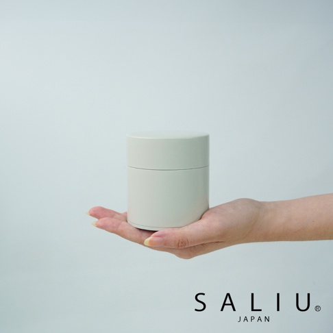 一個簡約的米白色圓柱形錫製茶葉罐，帶有蓋子，放在一隻手的手掌上。