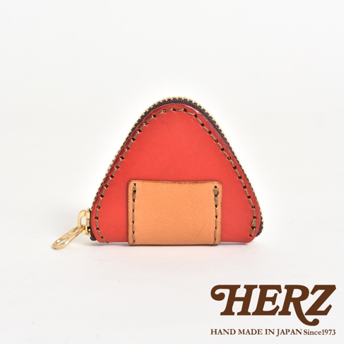 一個日本HERZ品牌的手作皮革三角飯糰造型零錢包，以紅色的軟皮革為主體，搭配正中央的淺棕色皮質插槽，邊緣飾有顯眼的縫線細節，側邊設有金屬拉鍊，下方印有HERZ品牌名稱與產地標示。