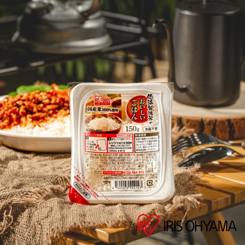 RIS FOODS 品牌標誌的低溫製法微波白飯，包裝上印有日文說明，標示「国産米100%使用」、「150g」、「低溫製法」等資訊。產品包裝為白色塑膠盒，部分透明。背景為戶外野營場景，有餐盤、餐具和炊具。