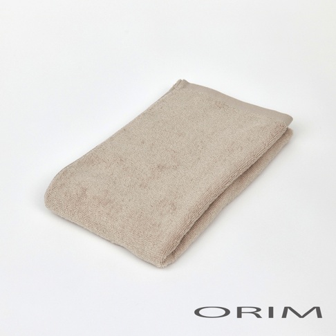 一條米色、柔軟的純棉迷你毛巾，織有細緻紋理，對折後放置於白色背景上，右下角有 ORIM 品牌標誌。