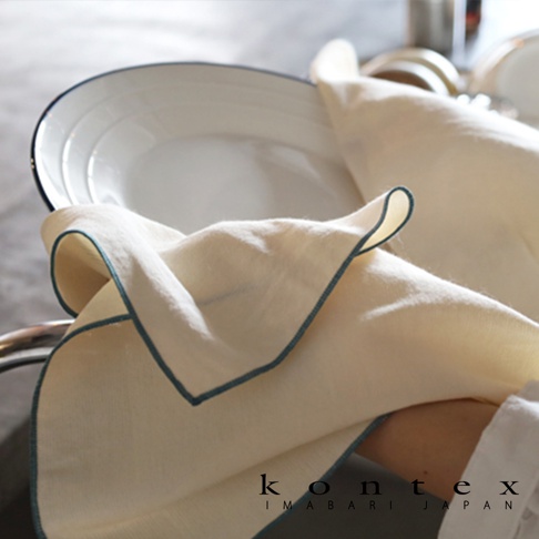 一張近距離拍攝的照片，畫面中一隻手正用一條乳白色的 KONTEX 今治和紙有機棉廚房拭巾擦拭白色盤子。拭巾邊緣綴有雅緻的深藍色滾邊，布料質感細膩輕盈，整體呈現出清新簡約的生活居家氛圍，下方並印有 KONTEX IMABARI JAPAN 的品牌標誌。