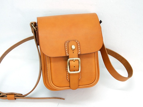 職人手作皮革皮扣直式單肩包，以天然皮革製成，呈現溫潤的琥珀色。包款設計簡約，正面配有金色金屬皮扣和縫線細節，搭配可調節的長皮革背帶。