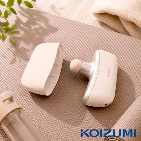 擺放在米色瑜珈墊上的 KOIZUMI 品牌筋膜槍，槍身為淺米色與銀色相間，機身保護蓋已拆卸放置於旁，露出圓形按摩頭，周邊另有透明水壺與折疊的瑜珈墊作為陪襯，整體氛圍簡約溫馨。
