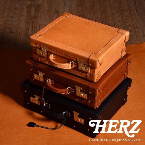 職人手作皮革英倫風便攜手提行李箱，堆疊擺放。最上方為淺棕色皮革，中間為中棕色皮革，最下方為黑色皮革。三只行李箱均有金屬鎖扣、皮革提把及金屬角飾。