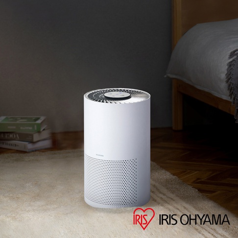 白色圓筒形空氣清淨機，底部有密集的圓形孔洞，頂部有扇葉和控制面板，置於淺色地毯上，背景為床鋪和書本。