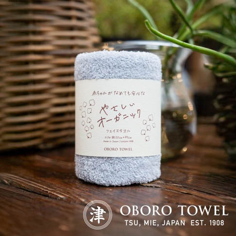 一條淺藍色的有機棉毛巾，質地柔軟，上面纏繞著印有日文說明和可愛圖案的白色標籤，標籤上註明尺寸為 33cm x 85cm，材質為 100% 棉。