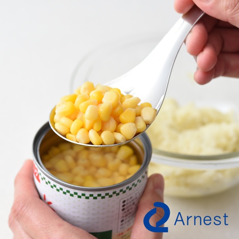 逸品物創不鏽鋼瀝水湯匙，銀色，盛著黃色玉米粒，湯匙邊緣有細小的瀝水孔，背景是裝有馬鈴薯泥的玻璃碗。