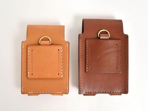 兩款職人手作皮革手機收納包，一款為淺棕色，另一款為深棕色。皆為直立式設計，正面有方形皮革片，上方連接黃銅 D 型環，側邊與底部有縫線裝飾。