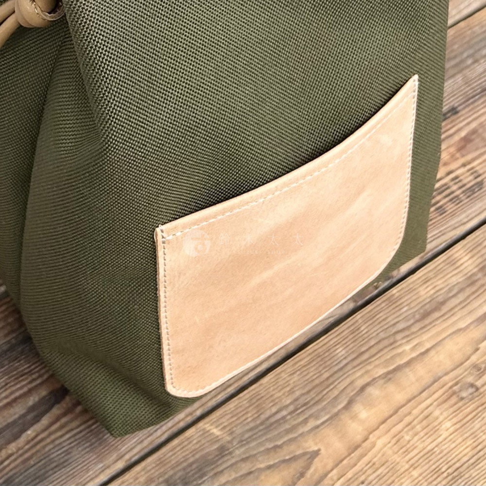 咖啡色皮革口袋縫製在軍綠色尼龍束口袋肩背包上，背景是木紋地板。