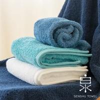 堆疊著三條不同顏色的浴巾：深藍色、藍綠色和白色。浴巾呈現柔軟的長纖維質感，其中藍色浴巾捲成一卷，另外兩條則對折堆疊。背景為柔和的米色。