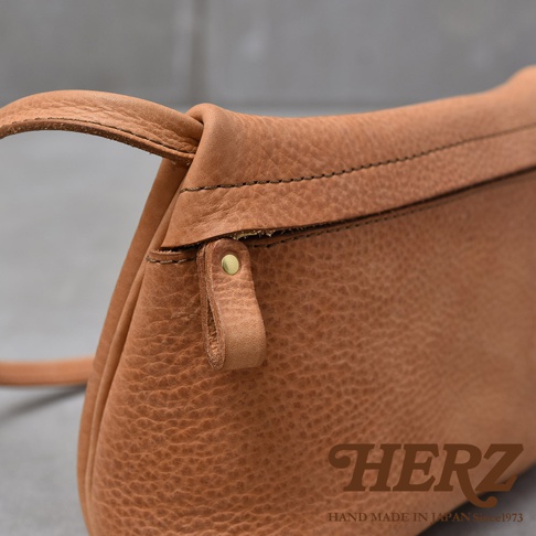 職人手作的淺棕色皮革斜背包，特寫拉鍊頭和皮革紋理，下方印有HERZ品牌字樣及HAND MADE IN JAPAN Since1973。