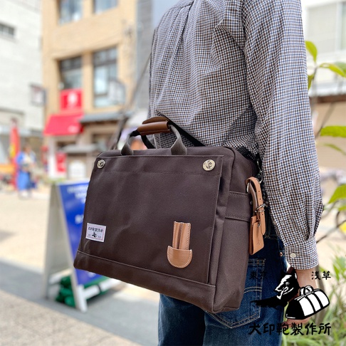 深棕色純棉帆布製成的B4商務公事包，配有皮革提把與裝飾。包款外側有一個翻蓋口袋，上面有皮革裝飾片，側邊掛有皮革吊牌。