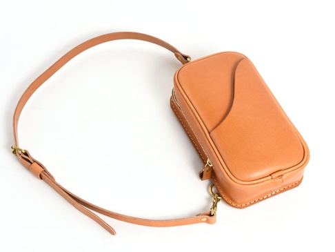 淺棕色皮革製成的直立型斜背包，帶有可調節的細長皮革肩帶和銀色金屬扣環，包身側邊有拉鍊開合，正面設計有弧形插袋。