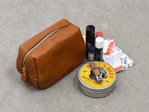 職人手作皮革Organ拉鍊收納包，淺棕色，光滑皮革材質，附金色拉鍊。旁邊散落著幾樣個人護理用品，包括藥丸、小瓶裝噴霧及圓形金屬罐。