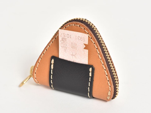 職人手作皮革零錢包，呈三角形，模仿飯糰造型。主體為淺棕色皮革，搭配黑色皮革裝飾，並縫有米色線跡。頂部設有銅色拉鍊，側面有一個金色金屬掛環。零錢包一角露出部分粉色紙張。
